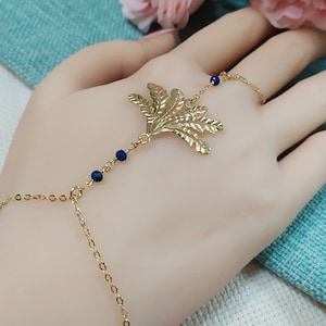 May include: A gold-coloured hand chain bracelet with a leaf-like design and small blue beads. The bracelet connects to a ring. The delicate piece is displayed on a hand, highlighting its intricate details and craftsmanship.