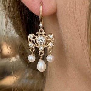 May include: Gold-toned chandelier earrings with a central clear gemstone and smaller clear stones. The earrings feature three dangling white pearls. The design is ornate and elegant, suitable for formal occasions.
