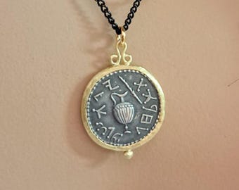 Puede incluir: Un collar con colgante de moneda plateada con bisel dorado y cadena negra. La moneda presenta un diseño en relieve y está suspendida de un cierre infinito dorado. El collar se lleva alrededor del cuello.