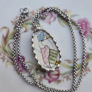 May include: A silver-toned necklace featuring an oval pendant with a scalloped edge. The pendant displays a hand-painted illustration of Jemima Puddle-Duck wearing a blue bonnet and pink shawl, with the text Jemima Puddle Duck clearly visible.