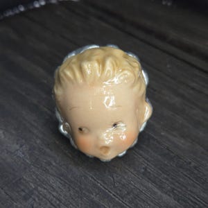 May include: A ceramic ring featuring a sculpted, pale-skinned face with blonde hair, rosy cheeks, and an open mouth expression. The face is set against a silver-toned, scalloped metal base that encircles the head, creating a detailed, dimensional accessory.