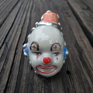 May include: A ceramic ring featuring a sculpted clown face with a white base, a bright red nose, red lips, and red accents around the eyes. The clown has a small orange hat on top and blue painted details on the sides, all set within a silver-toned metal band.
