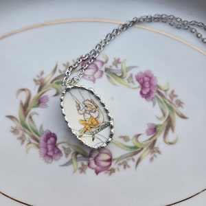 May include: An oval pendant necklace featuring a silver-toned scalloped frame and a central illustration of a frog wearing yellow trousers, sitting on a bench labeled J. FISHER, suspended from a matching silver-toned metal chain link necklace.