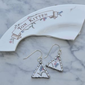May include: Triangle-shaped earrings with a floral pattern in pink and blue, set in a silver frame. The earrings hang from silver hooks. A broken ceramic piece with the text "Tom Kitten" is in the background.