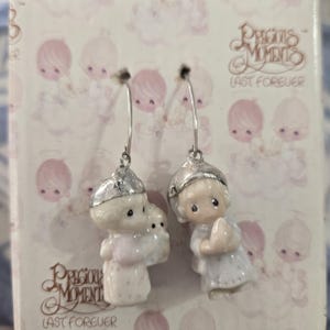 May include: A pair of dangle earrings featuring small, hand-painted porcelain figurines of children with teardrop-shaped eyes. Each figurine is topped with a silver-toned metal cap and attached to a silver-toned hook. The packaging displays the text Precious Moments Last Forever.