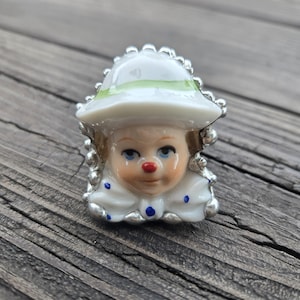 May include: A ceramic clown ring featuring a white hat with a light green band, a painted face with a red nose and blue eyes, and a white collar with blue polka dots. The ring band is silver-toned with a textured, beaded edge surrounding the face.