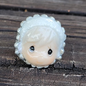 May include: A ring featuring a sculpted porcelain-style face of a child wearing a white bonnet with a scalloped edge. The face has a pale complexion, small nose, and two large, dark, teardrop-shaped eyes with a subtle, gentle expression.