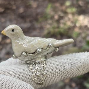May include: A ring featuring a small, glossy, off-white ceramic bird figurine mounted on a silver-toned metal base. The bird has a tiny black eye and a yellow beak. The ring band is made of ornate, filigree-style silver metal.