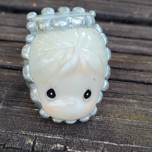 May include: A small, cream-colored ceramic figurine head featuring large, dark, teardrop-shaped eyes and a simple facial expression, framed by a textured, metallic silver border composed of small, rounded, bead-like elements.