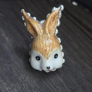 May include: A ring featuring a sculpted rabbit head with a tan textured fur pattern on the ears and forehead, a smooth white face, black eyes, and a small black nose. The band is made of silver-toned metal with a series of small, round beads.