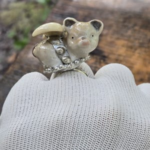May include: A ring featuring a small, cream-colored ceramic pig figurine with a glossy finish. The pig is adorned with a silver-toned band around its midsection, accented with small, round, metallic beads that encircle the base of the figure.