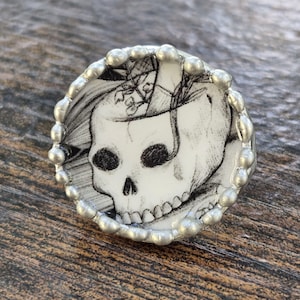 May include: A circular ring featuring a black and white ink illustration of a human skull with a plant growing from the top. The central image is encased in a silver-toned metal setting with a beaded, textured border around the edge.