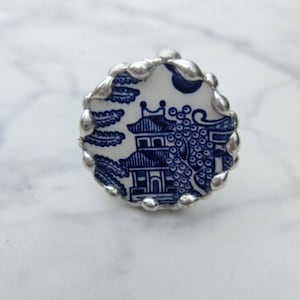 May include: A circular ring featuring a piece of white ceramic with a blue willow pattern, encased in a textured silver-toned metal setting with a scalloped edge. The design depicts a pagoda, trees, and foliage in a classic blue and white style.