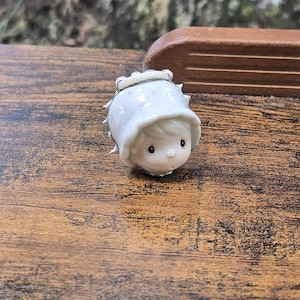 May include: A small, white ceramic figurine of a child wearing a bonnet. The figurine has a sweet, innocent expression with black dot eyes and a small, rosy mouth. The bonnet has a ruffled edge and is decorated with small pink dots. The figurine is set on a wooden surface.