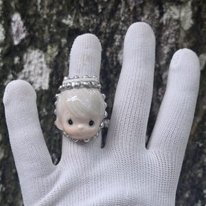 May include: A ring featuring a sculpted, pale-skinned child's face with large dark eyes and light hair, framed by a metallic silver border with small bead-like accents along the top edge, set on a band that encircles the finger.