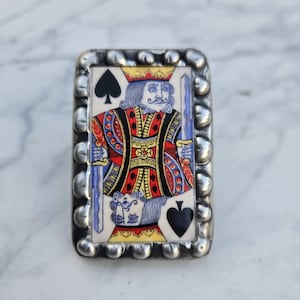 May include: A rectangular ring featuring a miniature illustration of the King of Spades playing card. The central image displays a royal figure in red, yellow, and blue attire holding a sword. The frame is made of silver-toned metal with a beaded edge.