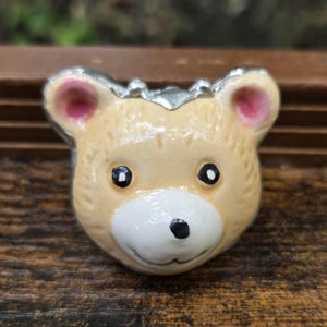 May include: A small, tan-colored bear-shaped brooch with a silver-colored trim. The bear has pink inner ears, black eyes, and a white muzzle with a black nose. The brooch is likely made of metal and enamel.