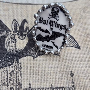 May include: An oval ring featuring a white background with black illustrations of a skull and crossbones, bats, and the text Bat Wings in venom. The ring has a silver-toned metal setting with a beaded edge surrounding the central design.