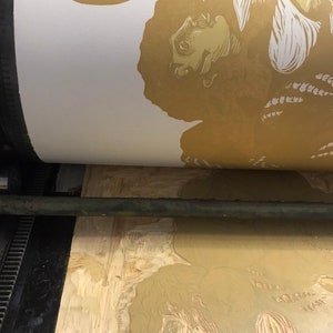 May include: A close-up of a printing press with a wooden block showing a carved image of a person's face and hair. The image is being printed onto a white sheet of paper.
