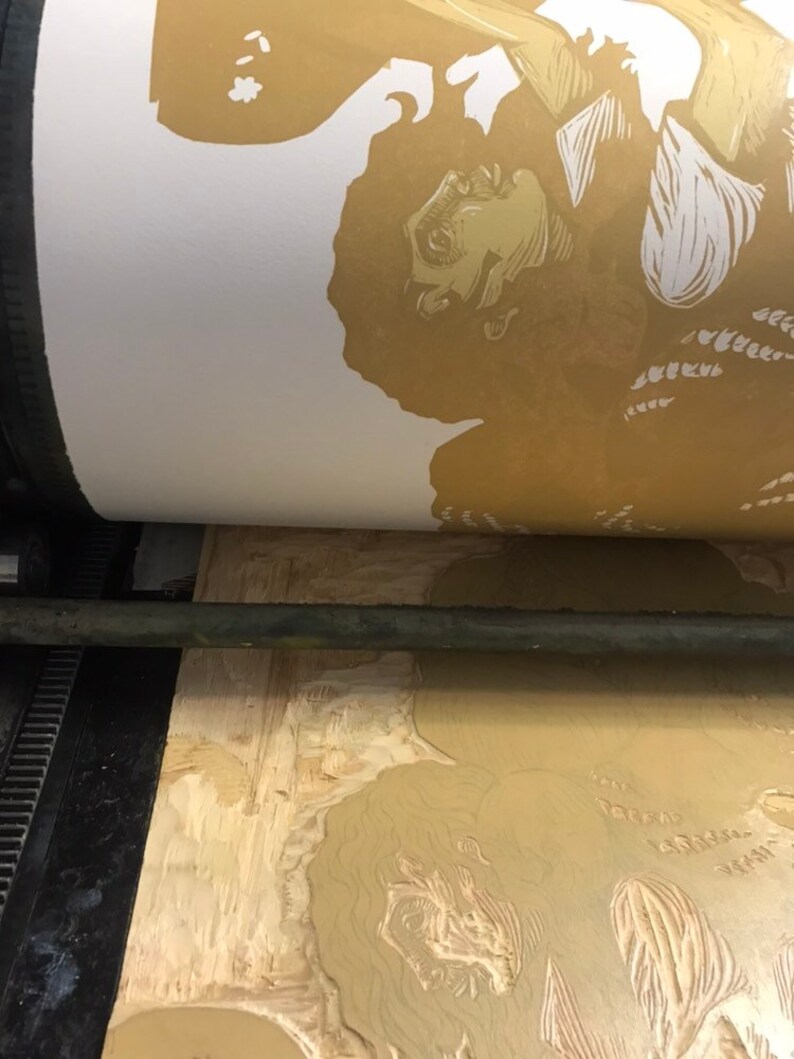 May include: A close-up of a printing press with a wooden block showing a carved image of a person's face and hair. The image is being printed onto a white sheet of paper.