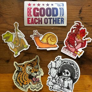 May include: Five colorful stickers featuring various illustrations. The stickers include a frog playing a banjo, a snail with a cowboy hat, a rooster playing a banjo, a tiger, and a skeleton wearing a sombrero and holding a cactus.