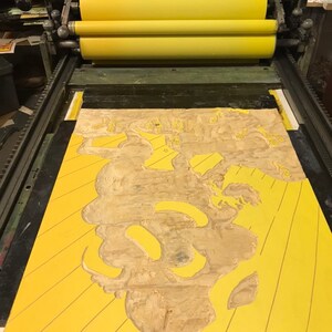 May include: A yellow printing press with a wooden block with a carved design on it. The block is being used to print a design onto a yellow surface.