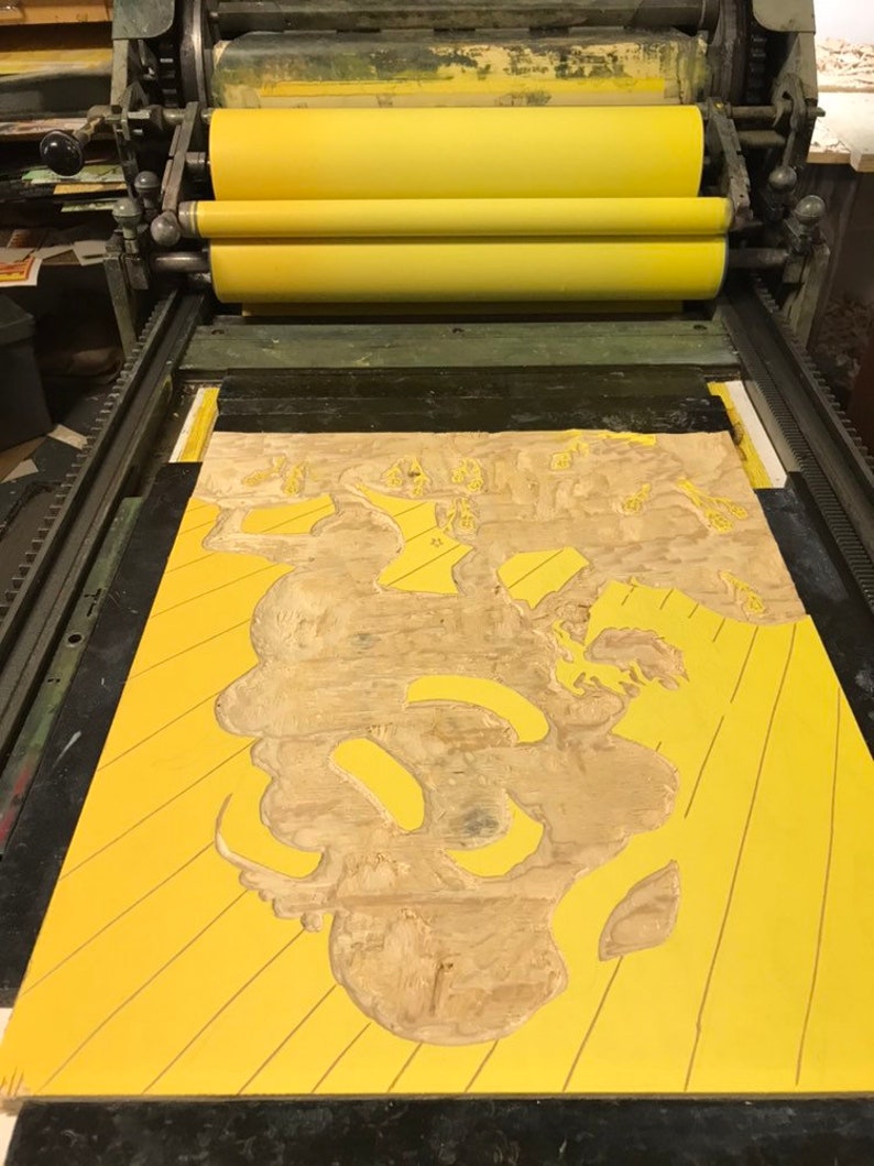May include: A yellow printing press with a wooden block with a carved design on it. The block is being used to print a design onto a yellow surface.