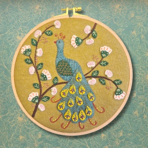 Op de afbeelding: Een ronde borduurring met een gele vilt achtergrond en een blauw en geel pauw ontwerp. De pauw is omringd door groene bladeren en witte bloemen.