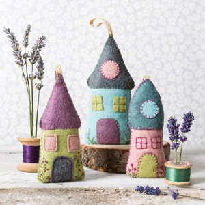 May include: Three felt houses with embroidered details, each with a different colour scheme. The houses are on a white surface with lavender sprigs in small vases.