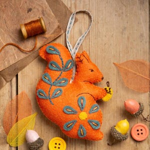 May include: Orange felt squirrel ornament with blue and green embroidered details, holding a yellow felt bee. The squirrel is hanging from a brown and white ribbon.