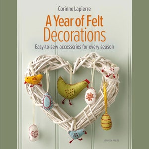 Op de afbeelding: Een boek getiteld "A Year of Felt Decorations" met een witte wilgenkrans in de vorm van een hart, versierd met viltversieringen, waaronder een geel kuiken, een groen kuiken, een blauwe vogel en verschillende eieren. De omslag van het boek bevat de tekst "Easy-to-sew accessories for every season".
