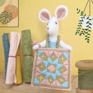 May include: A handcrafted white felt mouse figurine with pink-lined ears, wearing blue overalls, holding a small pink bird, and displaying a square quilt featuring a geometric pattern in shades of pink, blue, and olive green with yellow embroidery.