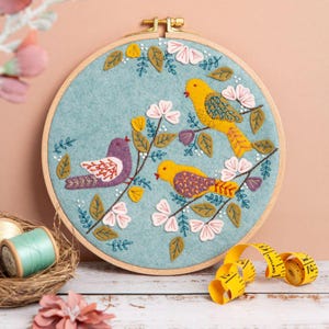 Op de afbeelding: Borduurwerk in een hoepel met drie gele vogels die op een tak zitten met roze en witte bloemen. De achtergrond is van lichtblauwe vilt.
