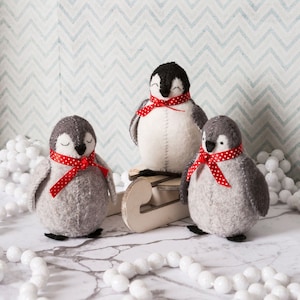 May include: Three grey felt penguins with red and white polka dot scarves. The penguins are sitting on a wooden sled. The background is a blue and white chevron pattern.