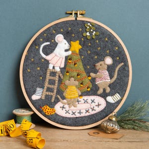 May include: A felt Christmas tree embroidery hoop featuring three mice decorating the tree. The mice are wearing pink, red and yellow outfits. The tree is green with brown acorns and a yellow star on top. The background is dark grey with white stars.