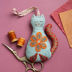 May include: A blue felt cat with a flower design on its chest and a long tail. The cat is lying on a pink surface with two spools of orange thread and a pair of gold scissors.