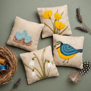 May include: Four square linen cushions with embroidered designs. The cushions display images of yellow flowers, a blue bird, blue eggs in a nest, and white snowdrop flowers. They are arranged on a green surface.