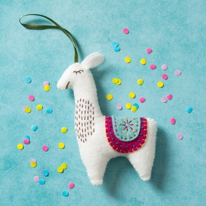 Op de afbeelding: Witte vilt lama-versiering met een blauw en roze geborduurd zadel. De lama hangt aan een groen touwtje en is omringd door kleurrijk confetti.