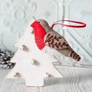Op de afbeelding: Een witte houten kerstboomversiering met een vilt roodborstje decoratie. De roodborst is rood, bruin en wit met geborduurde details. De boom heeft vijf kleine bellen.