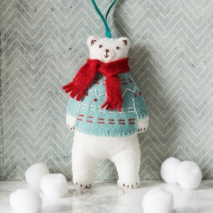Op de afbeelding: Een witte vilt-poolbeer ornament met een turquoise blauwe trui met rode stiksels en een rode sjaal. Het ornament is omringd door witte wattenbolletjes.