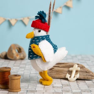 May include: A white felt seagull ornament wearing a red hat with a blue pom-pom, a teal blue scarf, and holding a yellow felt fish. The ornament is hanging from a string.