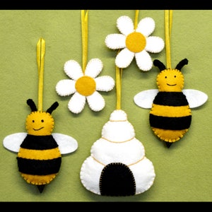 May include: A set of felt ornaments featuring a beehive, two bees, and two daisies. The ornaments are yellow, black, and white.