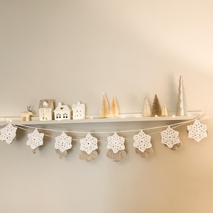 May include: A handmade crochet garland of white snowflakes with tan accents hangs below a shelf with miniature houses, trees, and a reindeer figurine. The garland is strung on a white cord, creating a festive, handmade decoration for the home.