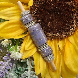 May include: A decorative lavender-colored bead and wood stick, adorned with silver and gold accents. The beads have a speckled design. The background features a yellow sunflower and lavender sprigs.