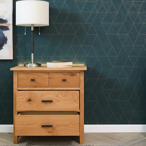 May include: A light brown wooden nightstand with three drawers, one open. A white lamp with a cylindrical shade sits on top, next to a stack of books. A tall green plant in a white pot is on the right.