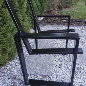 May include: Two black metal outdoor chairs with a minimalist design. The chairs have a rectangular frame and a backrest with a slight angle. The chairs are placed on a stone patio with greenery in the background.