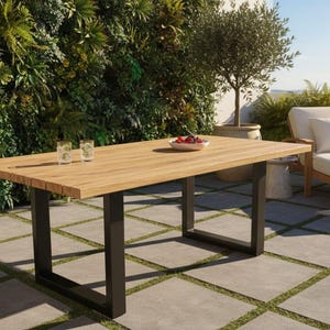 May include: A rectangular wooden outdoor dining table with a light brown finish and black metal legs. The table is set on a patio with a green wall in the background. A bowl of fruit and two glasses are on the table. A lounge chair is also visible.
