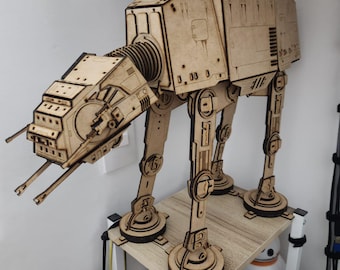May include: A wooden model of an AT-AT walker from Star Wars. The model is assembled and standing on a wooden shelf. The model is painted in a light brown color.