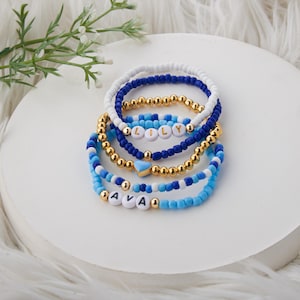 May include: A set of six beaded bracelets in shades of blue and white with gold accents. The bracelets feature the names "Lily" and "Ava" spelled out in white beads.