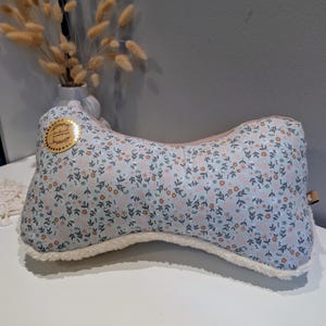 Op de afbeelding: Een lichtblauw, botvormig kussen met een bloemenpatroon in roze en wit. Het kussen heeft een zachte, witte, donzige rand en een klein, rond, goudkleurig label. Het kussen ligt op een wit oppervlak.